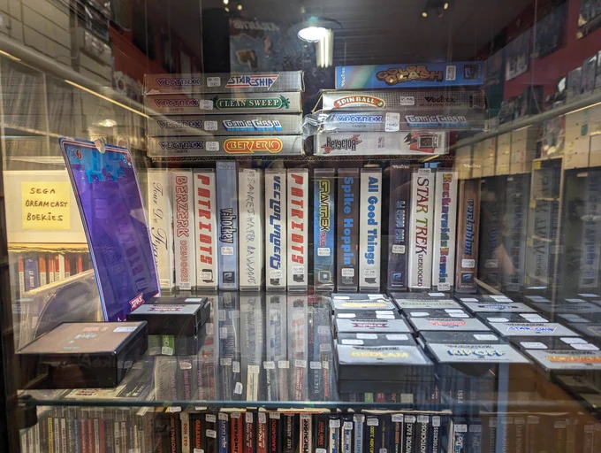 A glass case containing vectrex games. There are a number of assorted loose cartridges along the bottom and along the back row are the following homebrews: Tour De France, Berzerk Debugged, Patriots, v:hockey, Spike's Water Balloons, Patriots (second copy), Gravitrex plus, Spike Hoppin', All Good Things, 3D Space Cart, Star Trek Debugged, Rockaroids Remix, and Vectorblox. Stacked on top of those carts are the following boxed Vectrex games: Berzerk, Cosmic Crash, Clean Sweep, Star Ship, Color Clash, Spinball, Cosmic Chasm, and Revector. In the shelf below you can spot the spines of a number of Atari 2600 cartridges. 