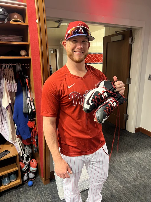 Photo of Craig Kimbrel holding a pair of his cleats in the clubhouse.  He is wearing the red and white pinstripe Phillies baseball pants, a red Phillies t-shirt and a red and blue Phillies hat.