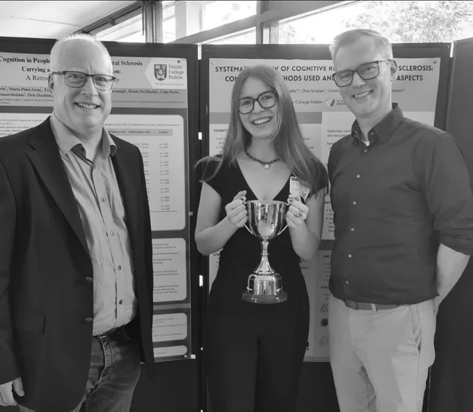 Clara Stein holding the DoN early career award perpetual trophy beside her research poster Systematic Review of Cognitive Reserve in MS: considering methods used and disease related aspects. She is standing with Kevin Twomey from Brainworx and Garret McDermott DoN chair