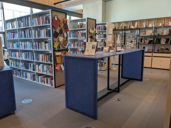 An image of the London College of Fashion Library. There is a long blue table with various materials placed across it. Behind are three shelves of books. In the background is a materials unit with various materials placed around it. 
