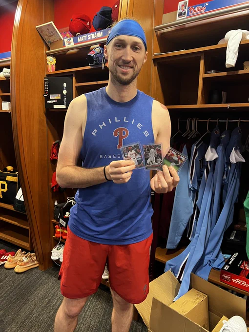 Photo of Matt Strahm in the Phillies clubhouse holding 3 baseball cards.  Strahm is smiling.  He's wearing a blue Phillies tank top, red shorts and a blue headband.