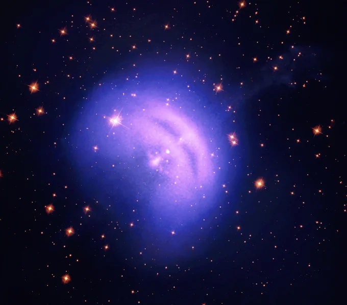 This image features the Vela Pulsar, the aftermath of a collapsed and exploded star sending a jet of particles into space. The pulsar resembles a soft, pillowy, lavender bean in a pocket of blue gas. A faint stream of gas, the X-ray jet, appears to shoot from the pocket, heading into the distance at our upper right. Purple markings in the lavender bean shape strongly resemble narrow eyes and an open mouth, giving the pulsar a squinting happy face. In this image, X-ray light is shown in blues and purples.
