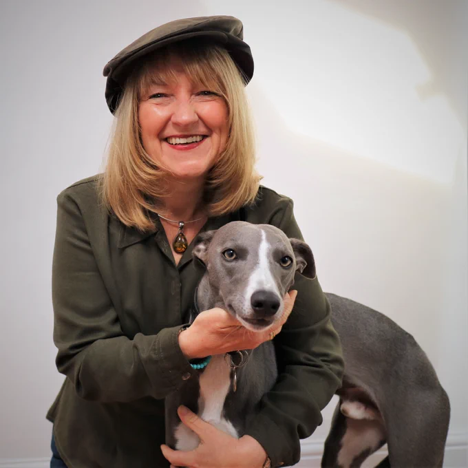 Sara Starling voice over smiling in flat cap & a dark green shirt, with a grey & white whippet snuggled under her arm.