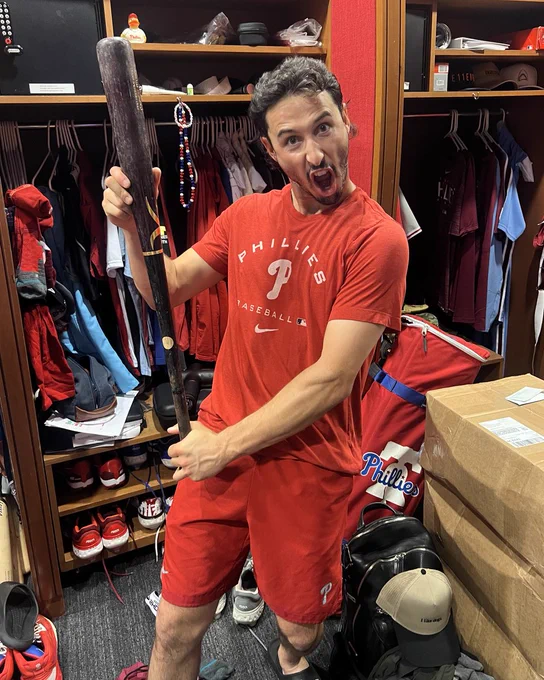 Photo of Garrett Stubbs holding one of his bats in the Phillies clubhouse.  He's making a funny face.  Stubbs is wearing a red Phillies t-shirt and red Phillies shorts.