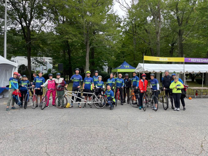 A group picture of our full team, most wearing Waypoint Adventure blue and green cycling jerseys, all yelling and smiling standing with their bikes on pavement. Behind them are refreshment tents and the Waypoint tent.