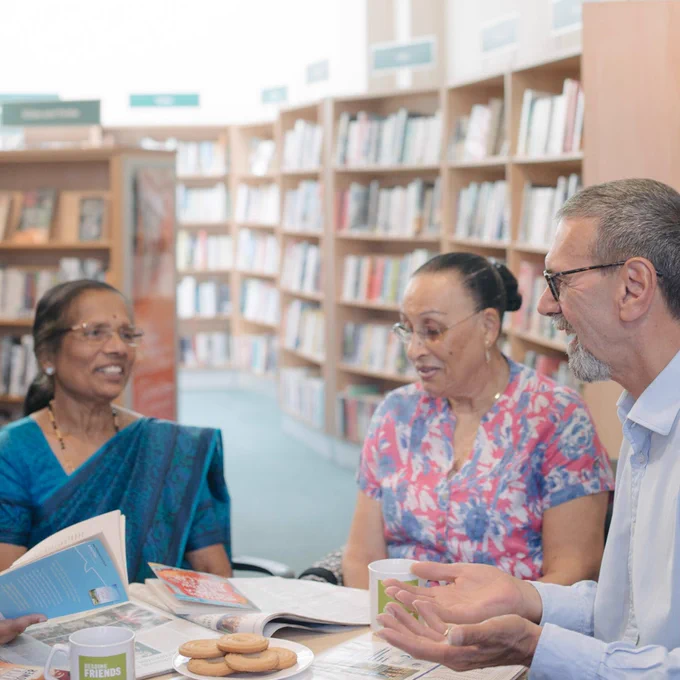 Three middle aged adults sat at a table in a library, talking. There are books and mugs on the table.