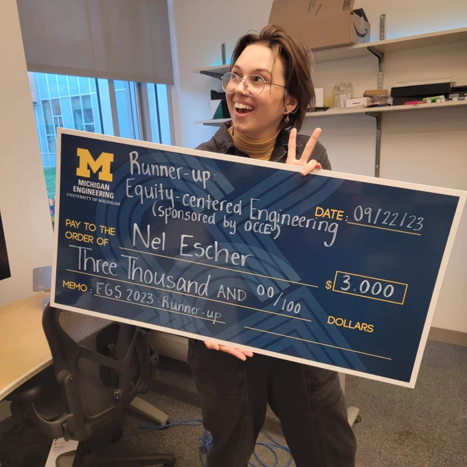 Nel holding an enlarged blue check showing the University of Michigan Engineering logo, the name of the award (Equity-centered Engineering), and a three thousand dollars amount.