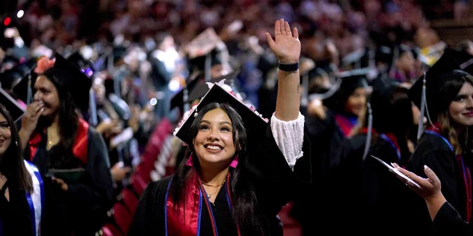 During commencement, a student in graduation regalia waves to someone in the crowd.