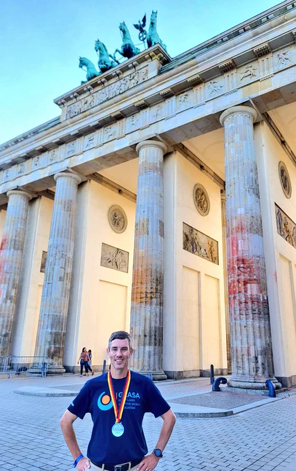 A man wearing a navy blue t-shirt for a charity GLASA (Great Lakes Adaptive Sports Association) with a finisher medal from the 49th Berlin Marathon stands in front of the Brandenburg Gate.