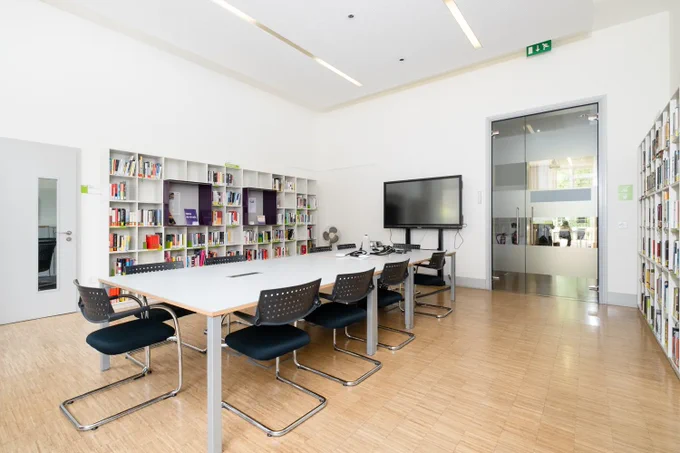 Interior photograph of a room with white walls, a wooden floor and a large glass door at the far end. The walls to the left and right have shelves filled with books, there's a large TV screen on the far wall, a large table surrounded by chairs in the centre and a door to the left.