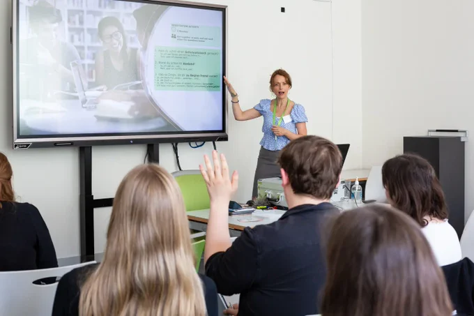 Photograph by Jayne Lloyd for Goethe-Institut London. Photograph in a classroom setting with a large screen on the wall to the left, a member of staff addressing a class whilst gesturing towards the screen and the backs of heads of people sitting in the foreground, one of which has their hand raised.