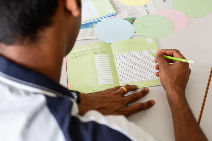 Photograph taken over the shoulder of a student who is working on a German wordsearch in a booklet, holding a green pencil in their right hand, with the back of their head to the left of the image. At the far end of the grey table they are working at is a newspaper and round pieces of coloured card with the letters 'D' and 'C' handwritten on them.