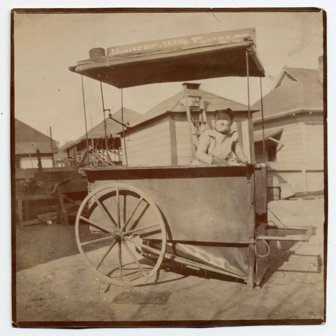 Alt Text: View of woman at two wheeled vending cart, sign on roof "Hunters Texas Tamales"; buildings in background; [Los Angeles, ca. 1897] 