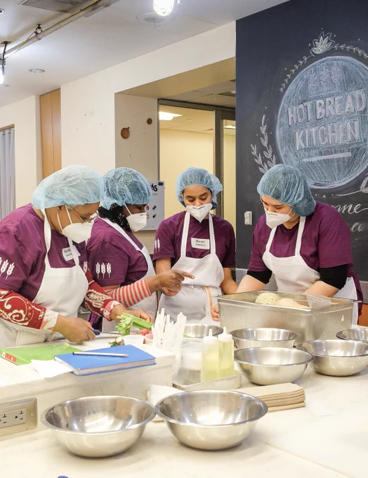 A team of workers in Hot Bread Kitchen cooking together. There are four women in purple shirts, white aprons, white masks, and blue hairnets around a table full of  bowls, as well as a few notebooks, and a small metal bin. Behind them to the right is a sign written on chalkboard wallpaper that says "Hot Bread Kitchen."