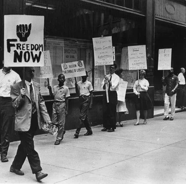 Black and white photograph of Black and white, middle-aged and young, people dressed very nicely walking in a line in front of a store. Each person holds a sign on a stick, the closest one has a closed Black fist and reads "Freedom Now."