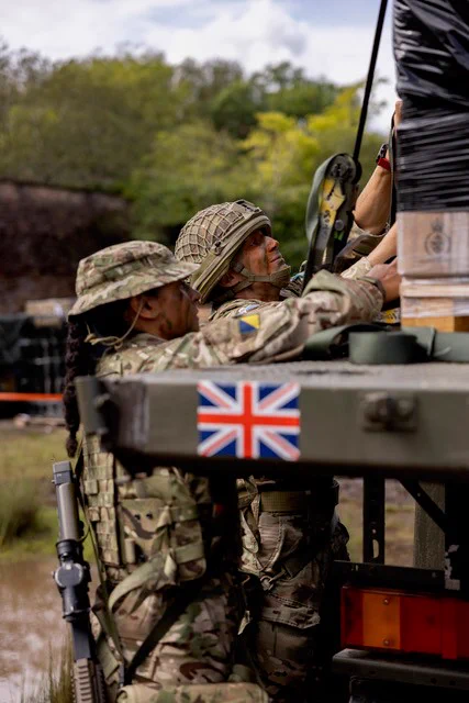 Two soldiers strap down cargo onto the trailer of a truck.