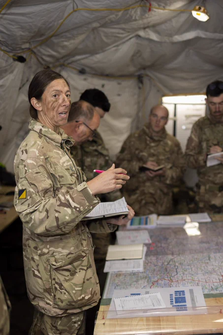 A soldier briefs her colleagues surrounded by maps and documents.