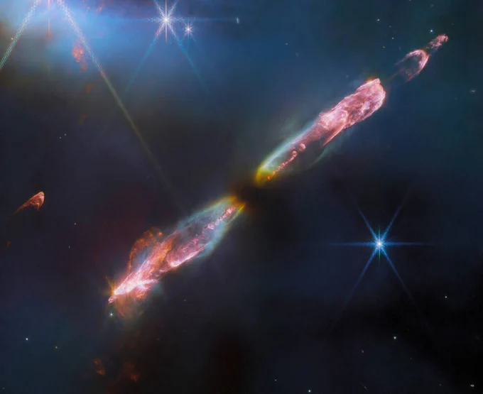 At the center is a thin horizontal pinkish cloud known as Herbig-Haro 211 that is uneven with rounded ends, and tilted from bottom left to top right. It takes up about two-thirds of the length of this angle, but is thinner and longer at the opposite angle. At its center is a dark spot. On either side of the dark spot, there are orangish yellow wisps that extend to light blue wisps. Within the center of those clouds, a pink fluffy streak runs through each lobe. At the ends of each lobe, pink becomes the dominant color. The lobe to the left is fatter. The right lobe is thinner, and ends in a smaller pink semi-circle. Just off the edge of this lobe is a slightly smaller pink semicircle, then a pink sponge-like blog. The background contains several bright stars, each with eight diffraction spikes extending out from the central bright point. Credits: ESA/Webb, NASA, CSA, T. Ray (Dublin Institute for Advanced Studies)