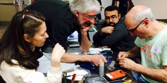 A photograph of four people at Brampton Library's Repair Cafe, one of whom is fixing an electronic device while the others watch. A variety of tools surround the fixer.  Two Repair Cafe participants are in the background.  