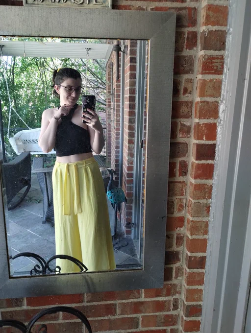 A mirror selfie of me, a white woman with short brown hair and glasses, on my porch. I'm wearing flowing, pale yellow pants and a sleeveless black crop top that crosses at the neck. I have my hair back in a yellow scrunchie and you can't really see it but I have a new gold nose ring!