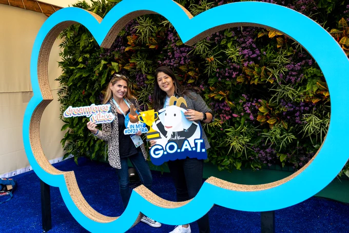 Two Dreamforce attendees taking a photo with a "Dreamforce Vibes" and "G.O.A.T." sign. 