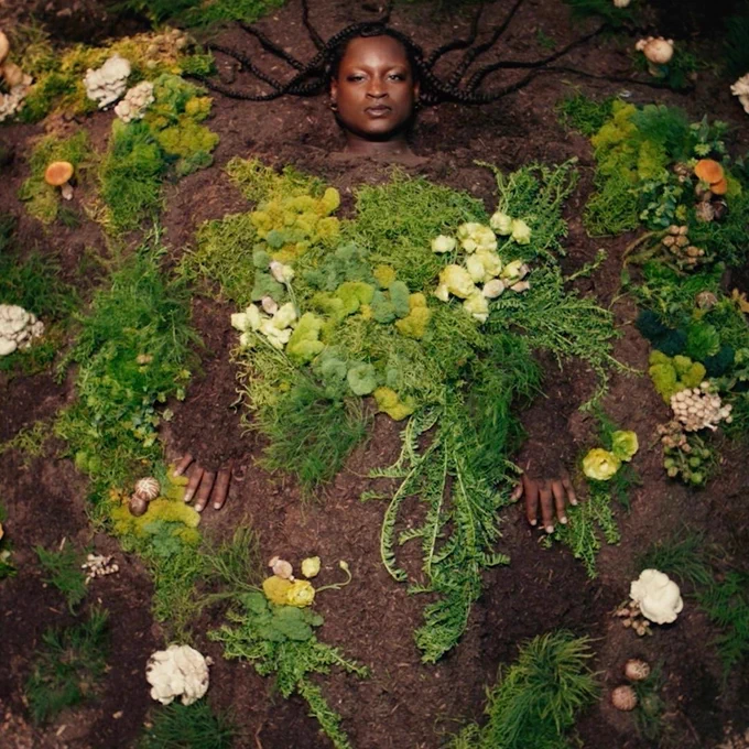 A black woman with braids lays underneath a garden of weeds, moss, and fungi.