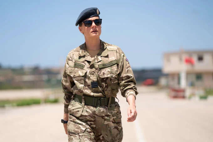 Eliza Russell (Romola Garai) walks confidently in the sun at what looks like an air base. She wears a camouflage shirt and trouser uniform, a beret and sunglasses.