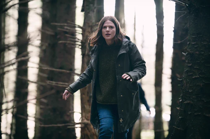 Kirsten Longacre (Rose Leslie) walks through a wooded area in coat and jeans, looking tense. Another figure is just visible behind her.