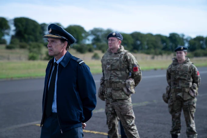 Marcus Grainger (Dougray Scott) stands in a dark navy coloured uniform, with a jacket, shirt and tie and a peaked pilot's style cap. Two soldiers in camouflage uniforms, sporting berets, stand behind him with their arms behind their backs.
