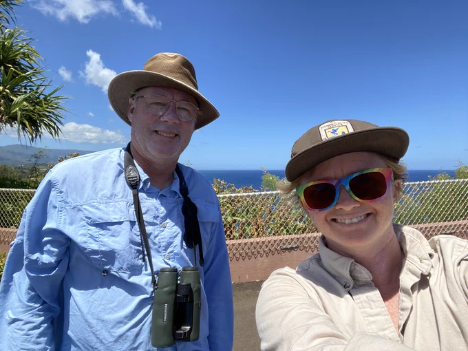 Two smiling people with brown hats pose for a photo, with the blue sky and ocean in the background.