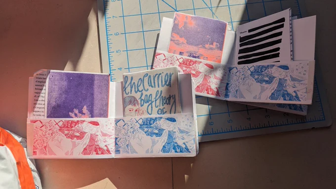A pair of zines printed in vibrant riso ink, featuring illustrations of Ursula Le Guin, shopping bags, and sunsets. Smaller zines are tucked into the pockets of the larger one
