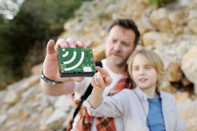 An adult and a child stand together in the background as the adult holds a small, green, rectangular device at arms length. The device is the focus in the foreground of the image. They both look as the child points at the device, which they will use to capture sound data during the upcoming annular solar eclipse as volunteers for the Eclipse Soundscapes Citizen Science project. 
