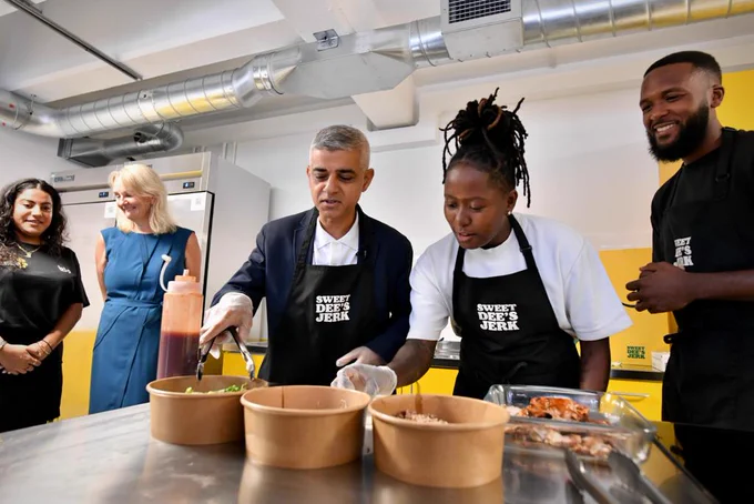 Mayor of London, Sadiq Khan wears an apron and serves food with another person, whilst three people look on. 