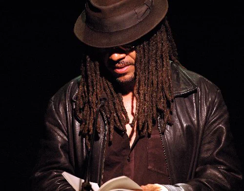 A Black man with dreads wearing a brown hat, brown leather jacket, brown shirt, and brown sunglasses looking down and reading a book