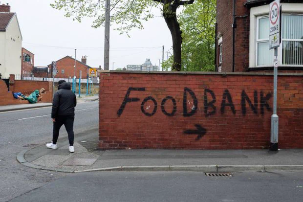 A man in a thick hooded jacket walks past a red-brick wall in which has been spray-painted the words “Food Bank” and an arrow pointing the way.