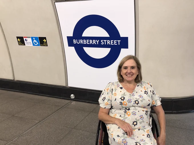 A picture of Tracey Proudlock in her wheelchair on the platform at Bond Street Station. The sign behind her says Burberry Street. She is smiling.