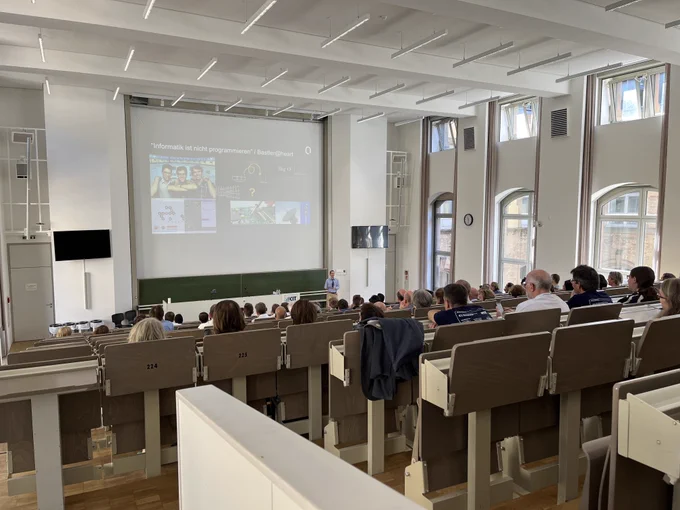 Bild eines Hörsaals mit Besuchern, die einem Vortragenden mit Folienpräsentation zuhören.