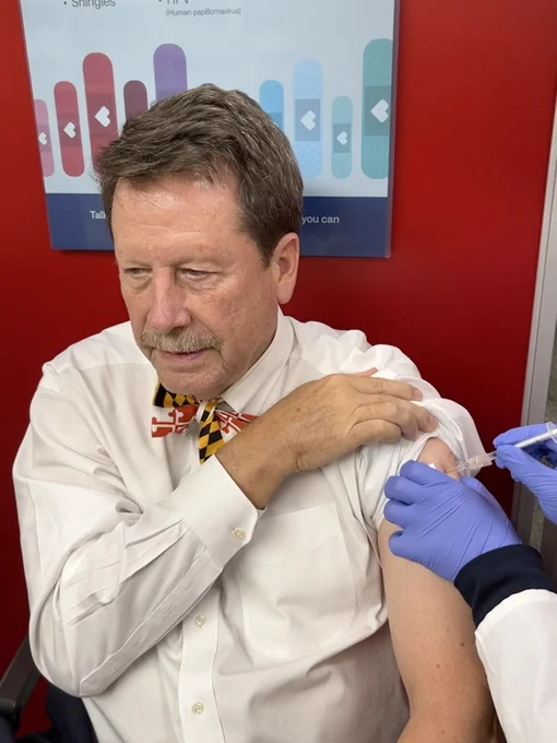 FDA Commissioner Dr. Robert M. Califf receives his COVID-19 vaccine in his left arm. He’s wearing a white shirt and a bow tie in the style of the Maryland state flag.