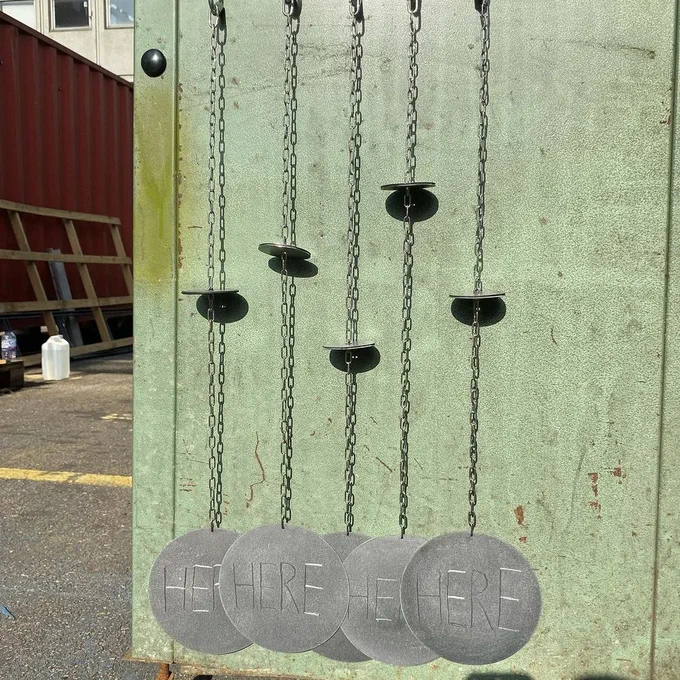 A photograph of 5 metal discs hanging on 5 chains in front of a green panel. Each disc has the word HERE in capital letters engraved in the middle of it. To the left of the green box we can see a car park flooring and a yellow line.

Image: © William Lang.