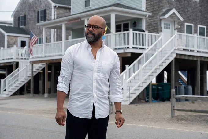 A film still from American Fiction, directed by Cord Jefferson. Jeffrey Wright is seen in a white button-up shirt and black pants standing in the middle of a street. Behind him are several two-storey beach homes and one has an American flag attached to its balcony. 