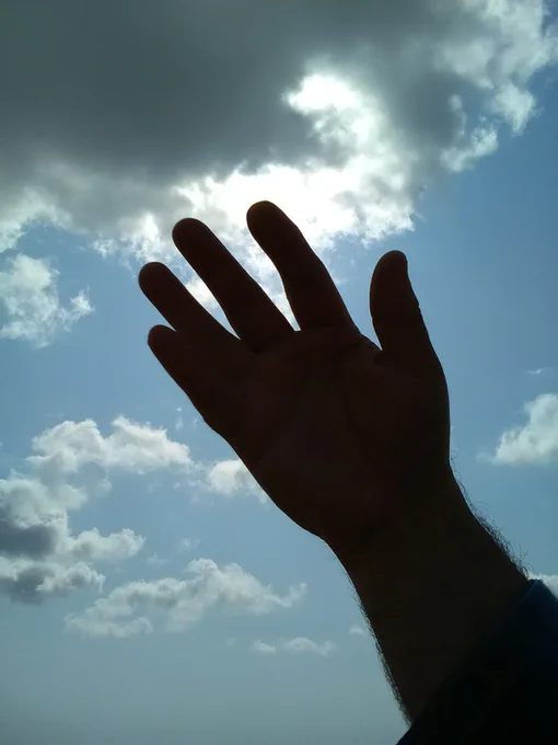 a silhouette of a hand, reaching up to a cloudy blue sky and the sun