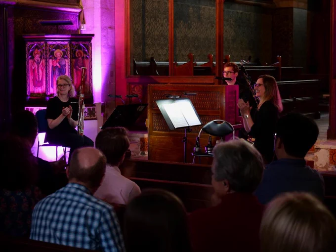 Three musicians from Britten Sinfonia taking applause from an audience at All Saints Cambridge church, with richly decorated painted walls in the background. 