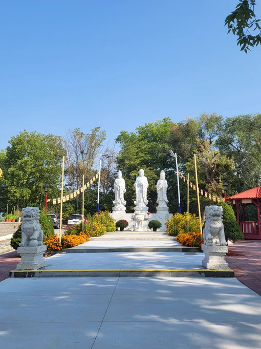Three white stone statues of Buddha stand in the back with a small walkway flanked by a pair of stone lions on pedestals. Behind the statues are several lush, green trees.