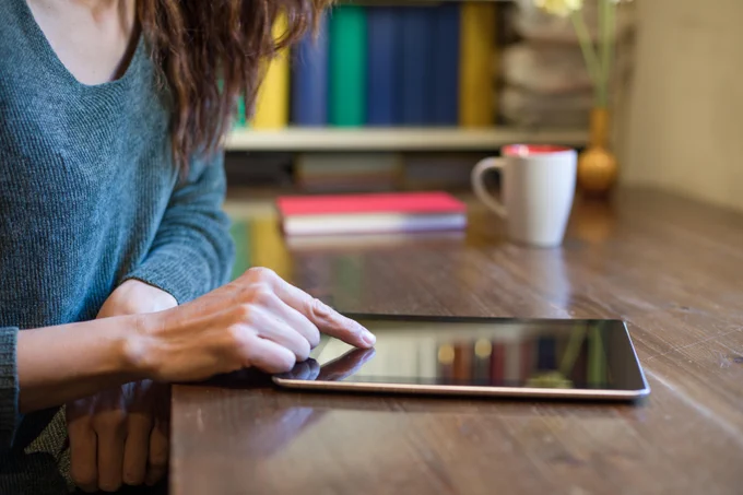 Photo of woman sitting at table using e-reader, with finger on screen. View focuses on e-reader – woman’s head out of shot.