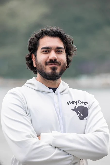 Nabeel Mansoor has curly dark brown hair and a beard and moustache. He is smiling slightly and has his arms folded. He is wearing a white hoodie with the HeyOrca logo, featuring a whale, on the left chest.