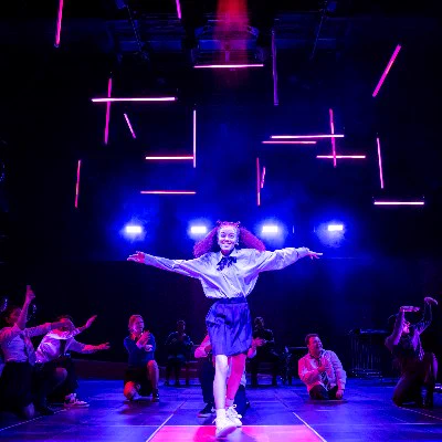A young woman stands in the middle of the stage in the midst of a dance off. Around her are young people crouched down and cheering her on. They wear white shirts, black skirts and/or trousers and striped ties. Behind them are pink neon lights shining down from thin lights, whilst four large blue spotlights shine from the back wall. 