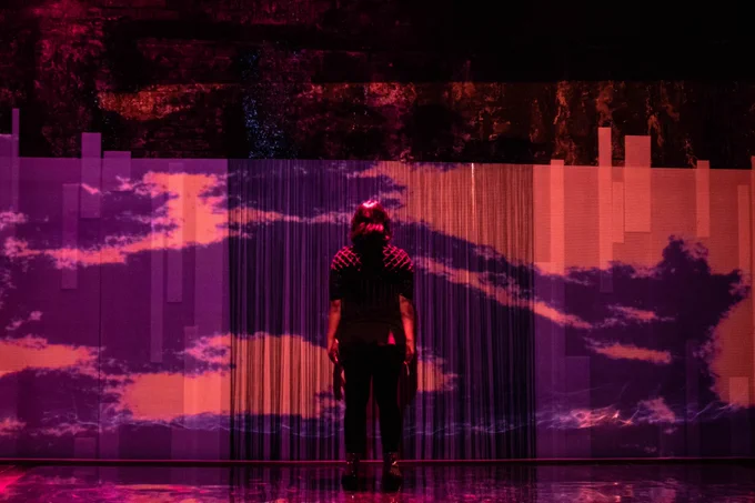 Peyvand Sadeghian performing on stage with their back to the audience. Purple and pink clouds are projected onto the back of the stage. Photo by Ali Wright.