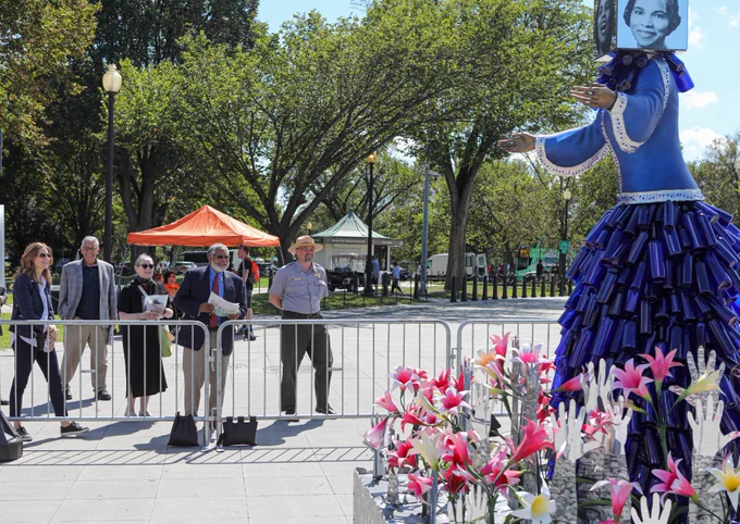 Five adults view a blue sculpture of Marian Anderson with outstretched arms, her skirt ringed by pink flowers. 