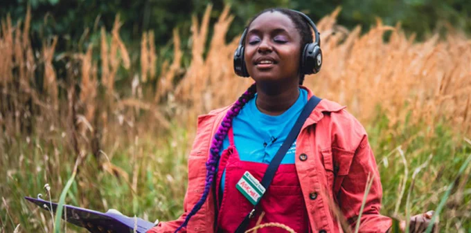 A young girl wearing headphones, a blue shirt, red dungarees and jacket stands in a field with her eyes closed. 