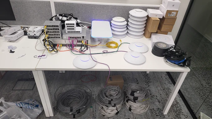 Bunch of Ubiquiti APs and switches laid on table, some powered on. Coils of network cables under the table. 
